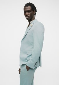 Άνδρας με πλεγμένα μαλλιά, φοράει γαλάζιο κοστούμι με καρφίτσα λευκό λουλούδι, στέκεται πλαγίως με το ένα χέρι στην τσέπη του.