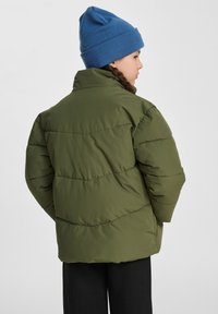 Olivgrön puffjacka med hög krage, med horisontell sömnad och avslappnad passform. Matchad med en blå stickad mössa.