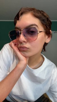 Grote, oversized zonnebril met gradient paarse glazen en gouden metalen accenten. Het montuur heeft een geometrisch ontwerp en ligt op een wit T-shirt.