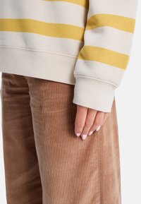 Beige gestreiftes Sweatshirt mit gelben horizontalen Linien; gerippter Bund. Braune Cordhosen mit vertikaler Textur. Hand ruht auf dem Bein.