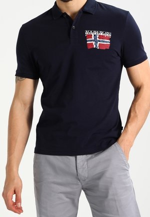 Mężczyzna w granatowej koszulce polo z norweską flagą i logo „Napapijri Geographic” na piersi, w połączeniu z jasnoszarymi spodniami.