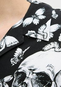 Crna tkanina s bijelim motivom lubanje, ukrašena detaljnim otiscima crnih i bijelih leptira. Upadljiva ovratnik s oštrim rubovima.