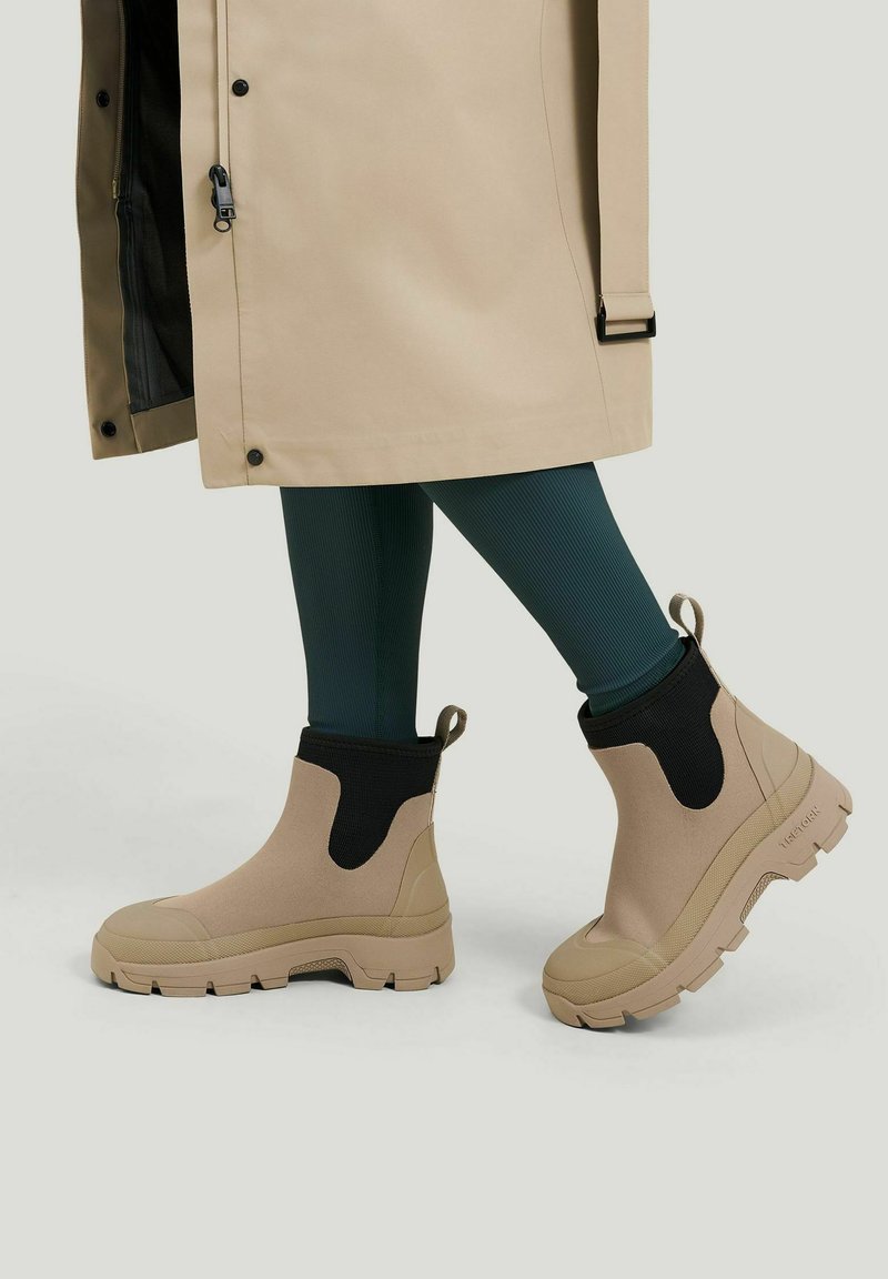 Beige chunky ankelstøvler med sorte elastiske sider, båret med mørkegrønne ribstrikkede leggings og en beige frakke med bælte og lynlåse detaljer.