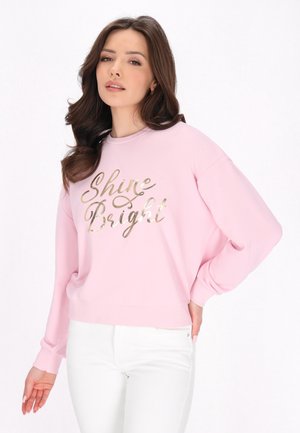 Felpa rosa con testo in foglia d'oro "Shine Bright." Presenta maniche lunghe, colletto girocollo e una vestibilità rilassata. Abbinata a pantaloni bianchi.