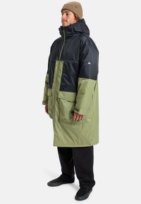 Long, padded coat with a dark navy upper and olive green lower. Features a high collar, zip closure, and large front pockets.