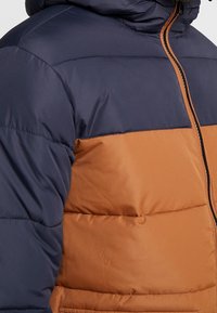 Kurtka pikowana z granatową górną częścią i brązową dolną częścią. Gładka faktura, zapinana na zamek błyskawiczny, z kapturem.