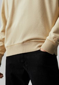 Sudadera de algodón beige con puños y cinturilla acanalados, que presenta un pequeño logo bordado en el puño, combinada con pantalones de mezclilla negros.