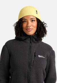 Frau mit lockigem Haar, die eine gelbe Strickmütze und eine schwarze, gefütterte Fleecejacke mit Reißverschluss und Jack Wolfskin-Logo trägt.