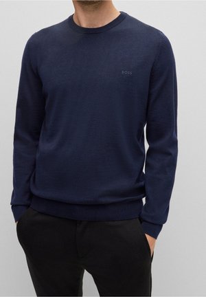 Homme portant un pull à manches longues bleu marine avec le logo "BOSS" sur la poitrine et un pantalon noir, mains dans les poches, fond neutre.