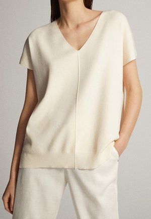 Vrouw draagt een crèmekleurige korte mouwen gebreide top met een V-hals en een naad in het midden, gecombineerd met bijpassende crèmekleurige broek, hand nonchalant in de zak.