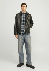 Chaqueta de cuero negra con cuello, superpuesta a una camisa de cuadros azul. El modelo lleva vaqueros de mezclilla claros y calzado negro.