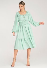 Miętowa sukienka midi z długimi bufiastymi rękawami, marszczonym wcięciem w talii oraz detalami z guzikami, wykonana z miękkiego materiału o warstwowym kroju.