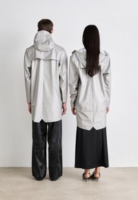 Manteaux imperméables gris avec capuches, design long et fentes à l'arrière, portés par-dessus des pantalons noirs et une jupe noire, vus de dos.
