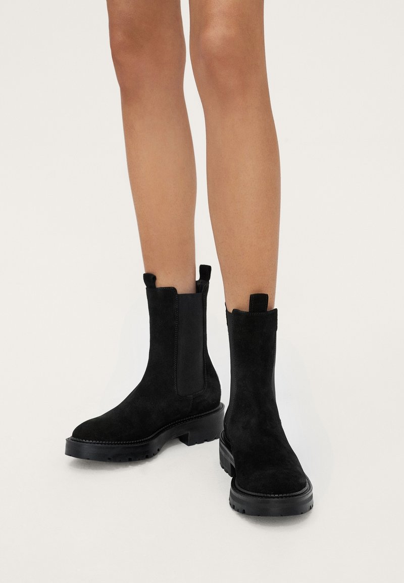 Bottines en suede noir avec des panneaux élastiques sur les côtés, semelle en caoutchouc plate et tirettes. Le design comprend une pointe arrondie et des détails de couture.