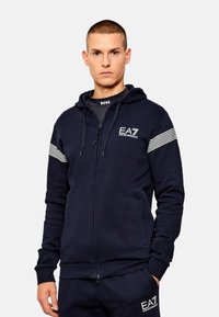 Felpa con cappuccio blu navy con chiusura a zip, tasca frontale e cordino sul cappuccio. Dettagli a righe bianche sulle maniche e logo EA7 sul petto. Tessuto dalla texture liscia.