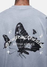 Jasnoniebieska bawełniana koszulka z dużym czarnym wzorem motyla oraz białym napisem: "NIEPRZYDATNY BRYTYJSKI ATELIER ZAŁOŻONE W 2011 R."