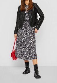 Schwarze Lederjacke über einem Maxikleid mit Blumenmuster in Marineblau und Weiß, kombiniert mit Stiefeletten und einer roten Umhängetasche.
