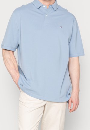 Homme portant un polo bleu clair à manches courtes et un pantalon couleur crème, une main dans la poche, sur un fond uni.