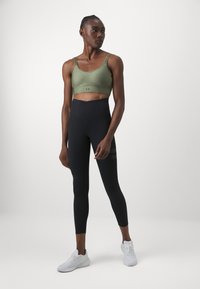 Bra desportivo verde com alças ajustáveis e textura leve, combinado com leggings pretas de cintura alta com um subtil detalhe riscado. Ténis brancos.
