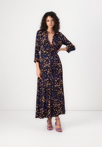 Maxi dress in navy with a colorful leaf print, featuring a collar, long sleeves, and button closure. Paired with purple strappy sandals.