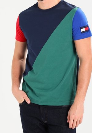 Homme portant un t-shirt à blocs de couleurs bleu marine, vert, rouge et bleu, avec un petit écusson rectangulaire représentant un drapeau sur la manche droite.