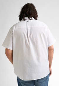 Witte short-sleeve shirt met een plooien aan de achterkant, ronde zoom en gestikte details bij de kraag. De stof lijkt lichtgewicht en textured.