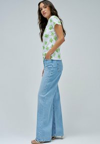 Camiseta blanca de manga corta con estampado floral verde, combinada con unos jeans de pierna ancha de color azul claro. Los jeans tienen un dobladillo deshilachado y un ajuste relajado.