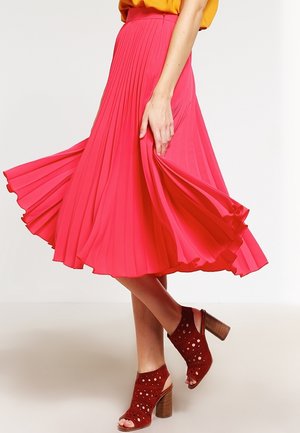 Femme portant une jupe midi plissée rose vif et des sandales à talons rouges perforées, tenant la jupe tout en étant debout.