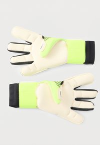 Par de guantes de portero con palmas beige, bordes de los dedos negros, correas de muñeca verde neón y puños negros, colocados planos sobre una superficie blanca.