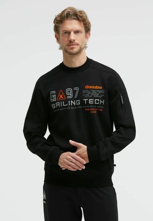 Man with curly hair and beard wears black sweatshirt with sailing-themed text and graphics, standing against plain light background.