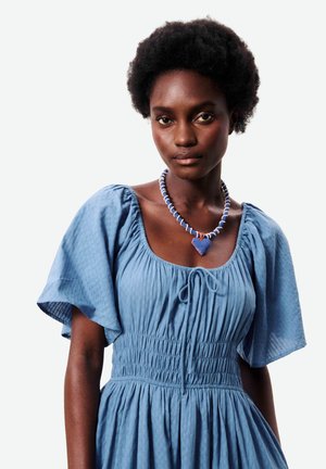 Vrouw met kort krullend haar, gekleed in een blauwe jurk met pofmouwen en een blauwe hartvormige kralenketting, staand tegen een effen achtergrond.