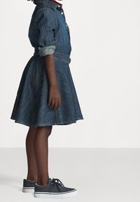 Jeansklänning med en figurnära överdel och utsvängd kjol, med uppvikta ärmar och en bälte i midjan. Matchas med mörka sneakers.