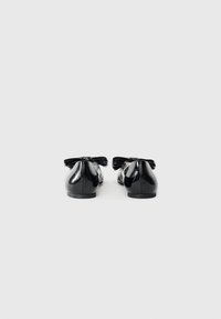 Ballerines en cuir verni noir avec un bout rond, ornées d'un grand nœud sur le talon et d'une texture lisse et brillante.