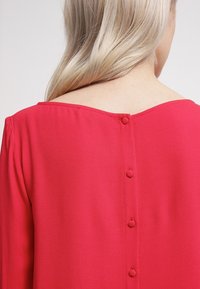 Blusa rosa brillante con una textura suave, que presenta una parte posterior baja y seis botones a juego cubiertos de tela a lo largo del cierre.