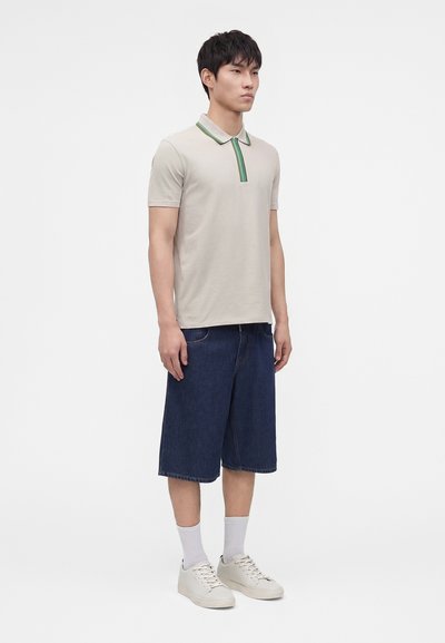 Homme debout portant un polo beige clair avec des détails verts, un short en jean foncé à longueur genou, des chaussettes blanches et des baskets blanches.