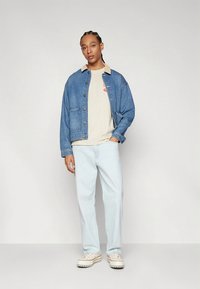 Denim jacket in medium blue with large front pockets, beige T-shirt featuring a logo, light blue jeans, and cream sneakers.
