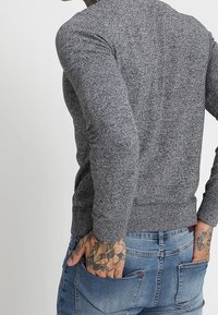 Grå, texturerad tröja med långa ärmar och en figurnära nederkant, bärs med blå denimjeans som har bakfickor.