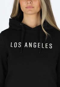 Black hoodie with a front print reading "LOS ANGELES" in uppercase white letters. Features a drawstring hood and a soft texture.