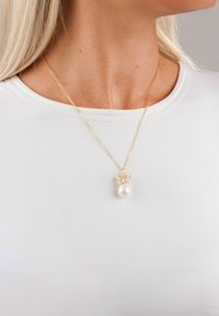 Guldkedja med en texturerad berlock, som har en enda vit pärla, bärs över en ljusfärgad t-shirt med rund hals.