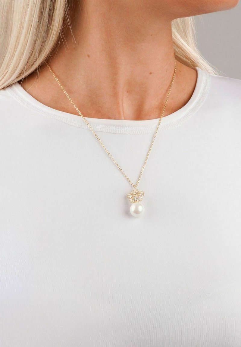 Guldkedja med en texturerad berlock, som har en enda vit pärla, bärs över en ljusfärgad t-shirt med rund hals.