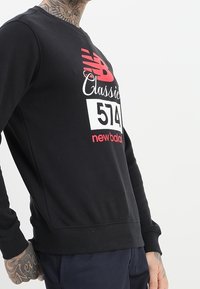 Felpa nera con maniche lunghe, con grafica rossa e bianca con le scritte "New Balance" e "Classic 574" sul davanti. Tessuto morbido.