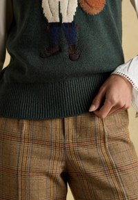 Tumši zaļš adīts džemperis ar grafisku dizainu krūšu daļā, apvienots ar brūnām svītrainām biksēm, kuras ir ar ziliem un sarkaniem svītrām.