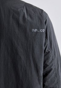 Grå jakke med en struktureret finish. Har et logo "NN. 07" trykt i lys grå på skulderen. Minimalistisk design, moderne silhuet.