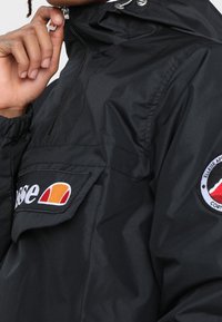 Sort nylon hættejakke med en frontlomme med lynlås, elastiske manchetter og et logo-patch i orange og hvid på brystet.