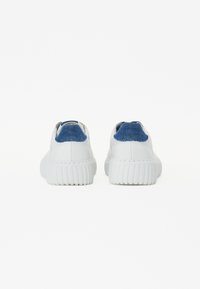 Adidași albi cu o talpă din cauciuc texturat. Spatele are un accent din denim albastru la călcâi, contrastând cu partea superioară netedă de culoare albă.