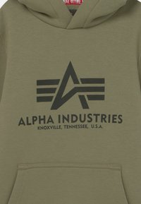 Olivově zelená mikina s kapucí vyrobená z měkké látky, zdobená velkým černým logem s křídly a textem "ALPHA INDUSTRIES, KNOXVILLE, TENNESSEE, U.S.A."