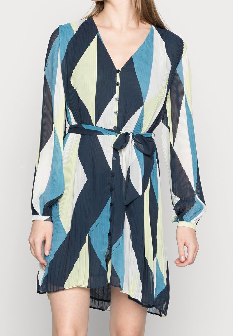 Femme portant une robe plissée à col en V avec manches longues, motif géométrique en bleu, noir, blanc et vert clair, et une ceinture nouée à la taille.