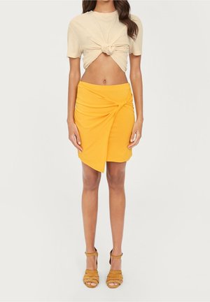 Top beige court avec un devant noué et une jupe enveloppante jaune avec un détail tordu. Les sandales ont un design à lanières en marron.