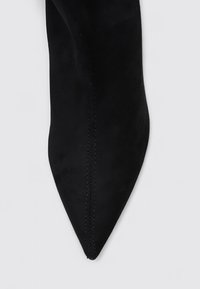 Zapato negro de punta afilada hecho de ante, con una textura suave y costuras visibles a lo largo de la costura, situado sobre un fondo claro.
