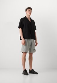 Camisa negra de manga corta con un patrón texturizado, combinada con pantalones cortos grises hasta la rodilla y zapatillas deportivas negras. El conjunto tiene un corte casual y relajado.